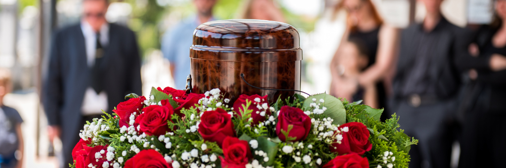 Picture of an urn containing ashes. Stood among red roses with mourners in the background.